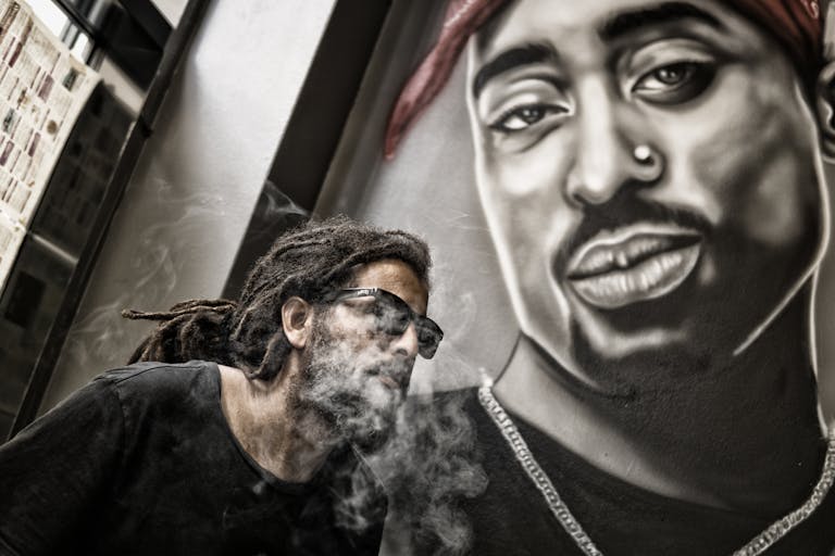 Man With Dreadlocks and Sunglasses Poses Near Tupac Shakur Portrait