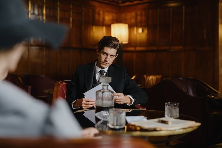 A Man in Black Suit Sitting Near the Table while Holding a Paper