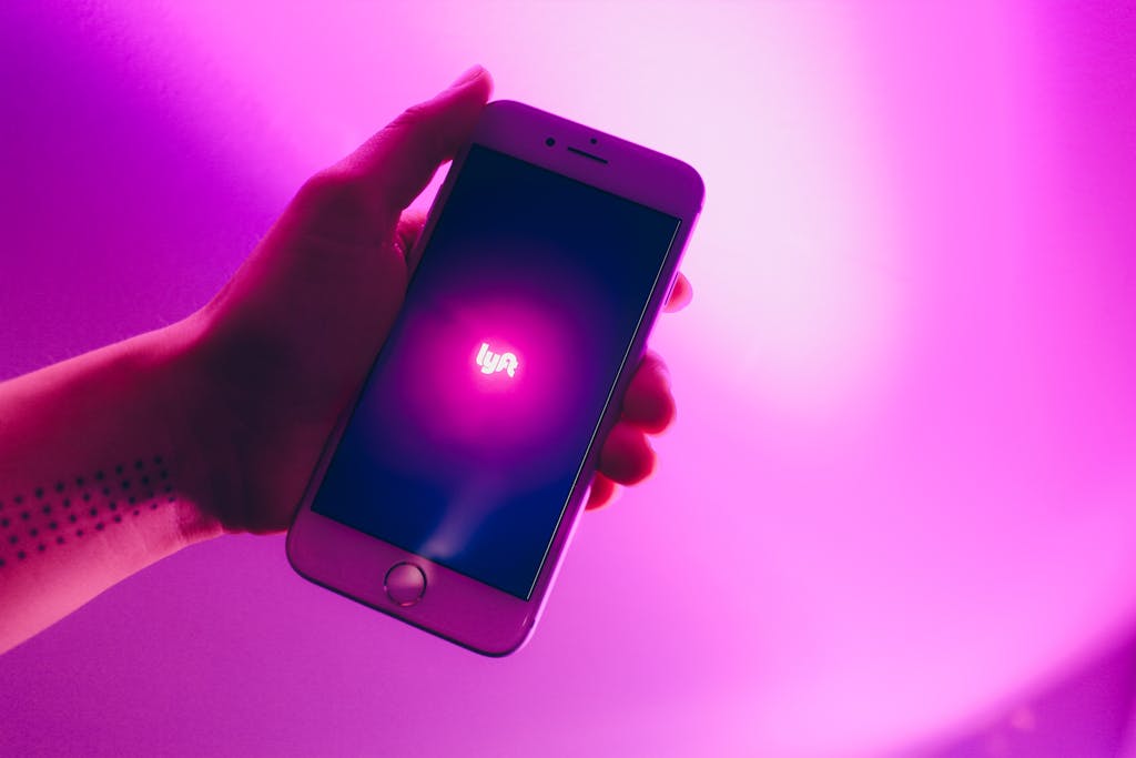 A hand holding a smartphone displaying the Lyft ridesharing app with a vivid pink background.