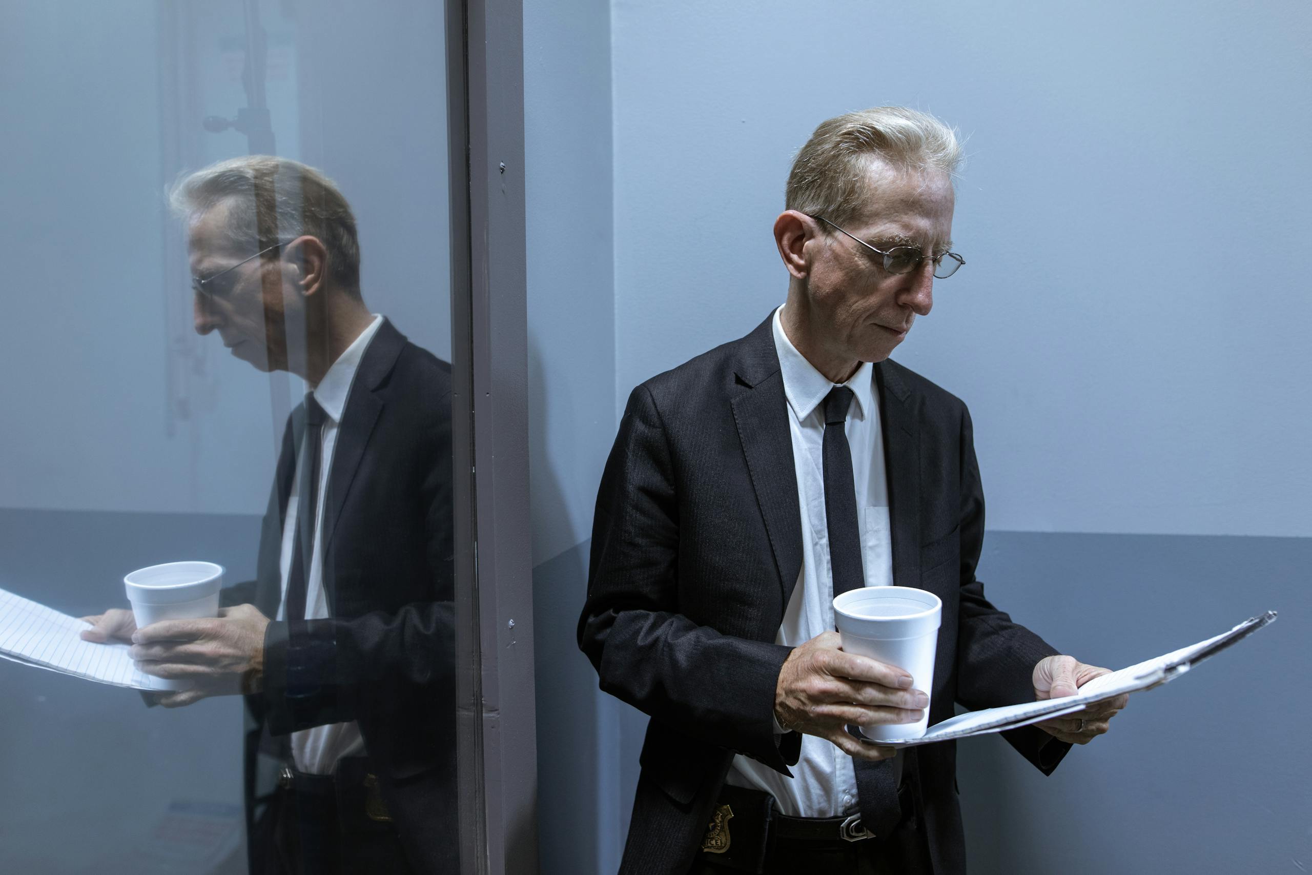 A businessman holding coffee, reviewing documents in an office hallway with reflection.