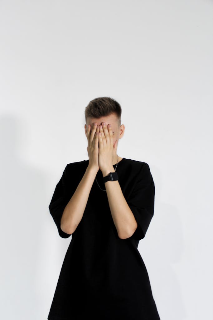 A man in a black shirt covers his face with hands in a studio shot against a white background.